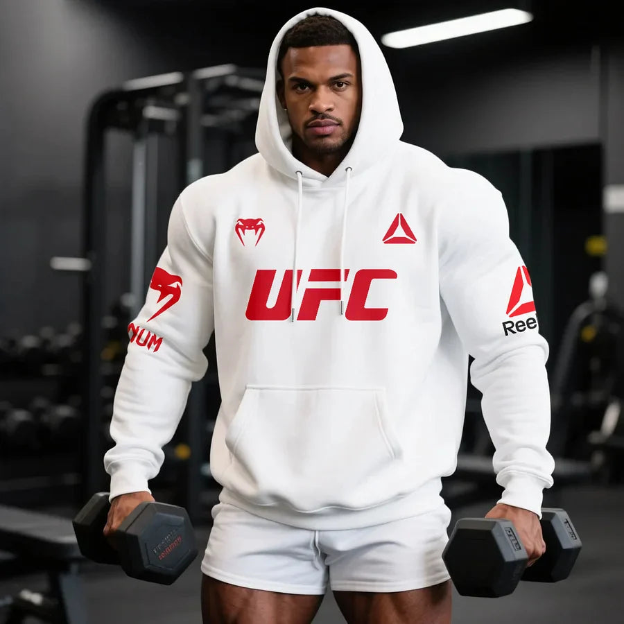 Mężczyzna atletyczny noszący biały hoodie VenuReebk z czerwonym logo UFC, na siłowni, trzymający hantle.
